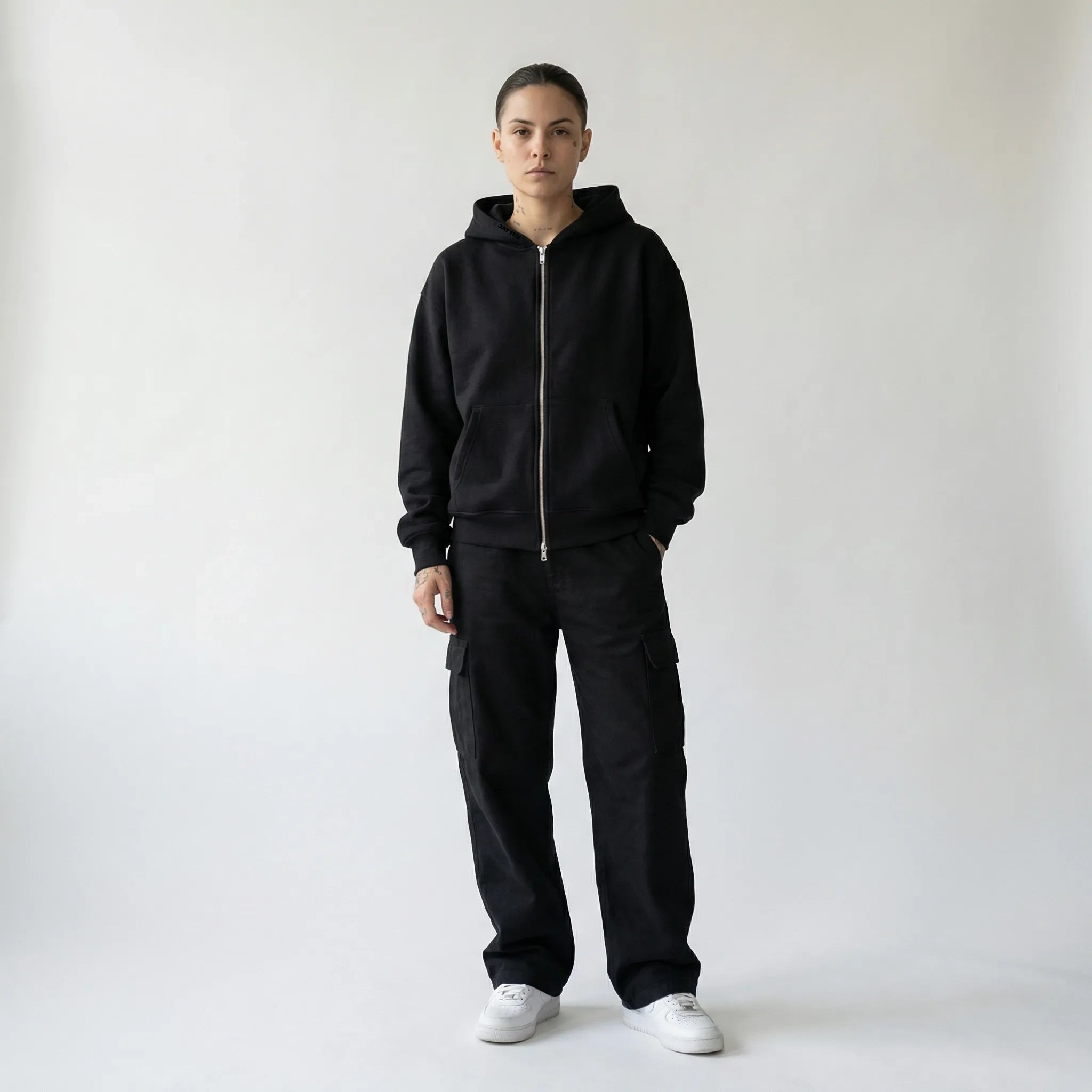 Person wearing a black JAYTEE hoodie and pants with white sneakers on a light gray background