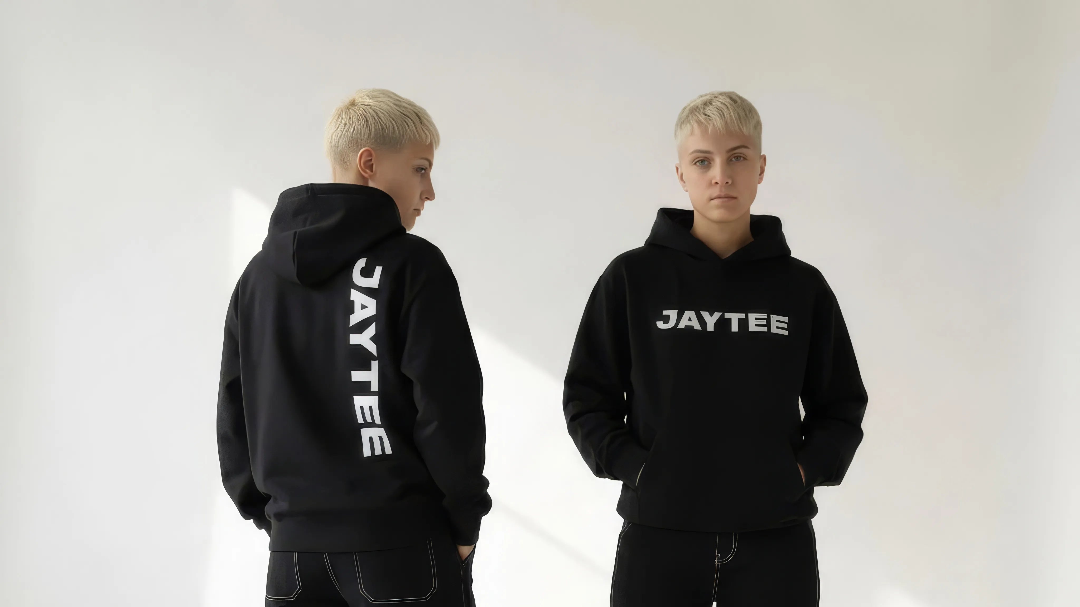 Two people wearing black 'JAYTEE' hoodies on a white background
