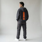 Person wearing a dark gray JAYTEE hoodie and pants with an orange exclamation mark on the back, standing against a plain background.