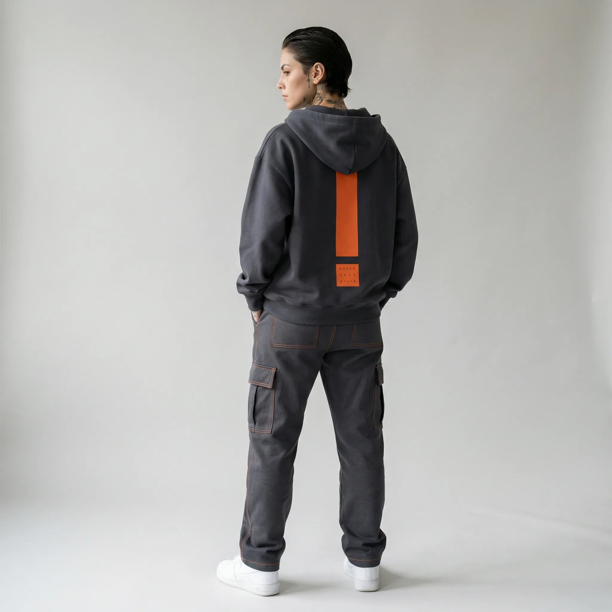 Person wearing a dark gray JAYTEE hoodie and pants with an orange exclamation mark on the back, standing against a plain background.