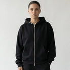 Person wearing a black JAYTEE hoodie and pants with white sneakers on a light gray background