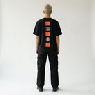 Person wearing a black JAYTEE t-shirt with orange graphic designs on the back, standing against a plain white background.