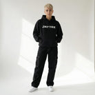 Person wearing a black JAYTEE hoodie with 'JAYTEE' branding and black cargo pants on a white background