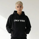 Person wearing a black JAYTEE hoodie with 'JAYTEE' branding and black cargo pants on a white background
